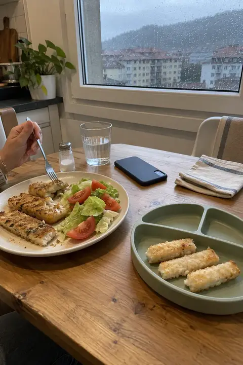 Mesa junto a una ventana con lluvia: un plato de adulto con palitos de merluza y ensalada y un plato de silicona con tres palitos de merluza para el bebé