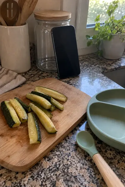 Móvil apoyado en un bote sobre la encimera, junto a una tabla con calabacín y un plato de silicona
